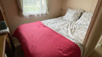 Un lit simple avec une couverture rouge, une fenêtre ornée de rideaux en dentelle et une tête de lit en bois.
