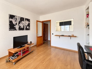 A modern living room with a flat-screen TV on a wooden stand, a small table with a cup, and a mirror above a window. - 2BR Tamarán House - Central & Quiet LPGC (Las Palmas de Gran Canaria)