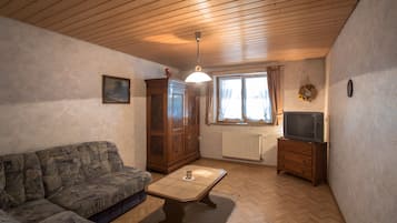 Ein Wohnzimmer mit Holzbalkendecke, einer Couch, einem Schrank, einem Fernseher auf einem Schrank und einem Fenster mit Vorhängen.