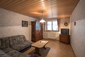 Ein Wohnzimmer mit Holzbalkendecke, einer Couch, einem Schrank, einem Fernseher auf einem Schrank und einem Fenster mit Vorhängen.