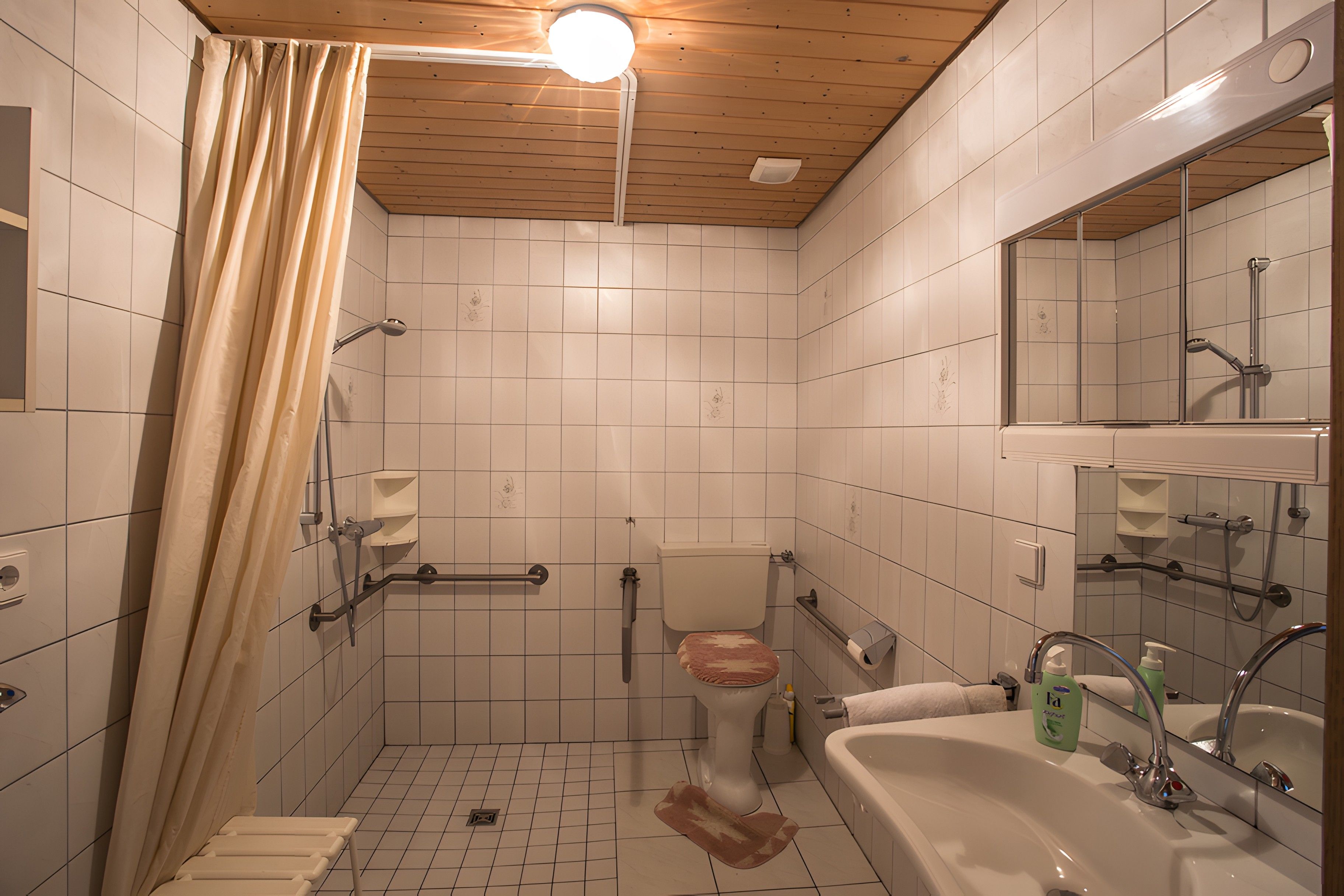 Ein Badezimmer mit weißen Fliesen, einer Holzbalkendecke, einer Duschkabine, einer Toilette, einem Waschbecken mit Spiegel und einem Handtrockner.
