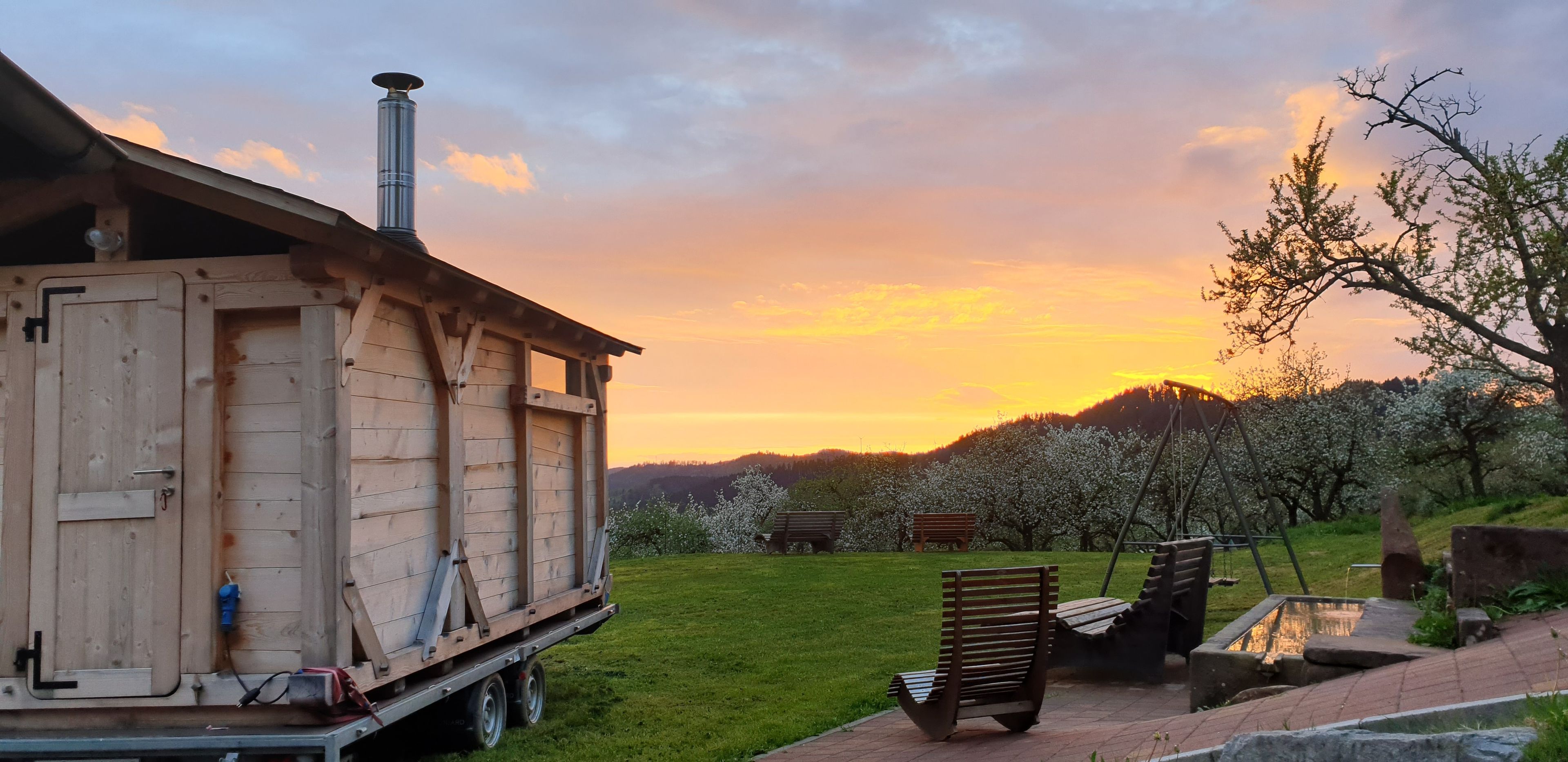A wooden structure with a chimney, a bench, and a swing set in a grassy area with trees and a sunset in the background.