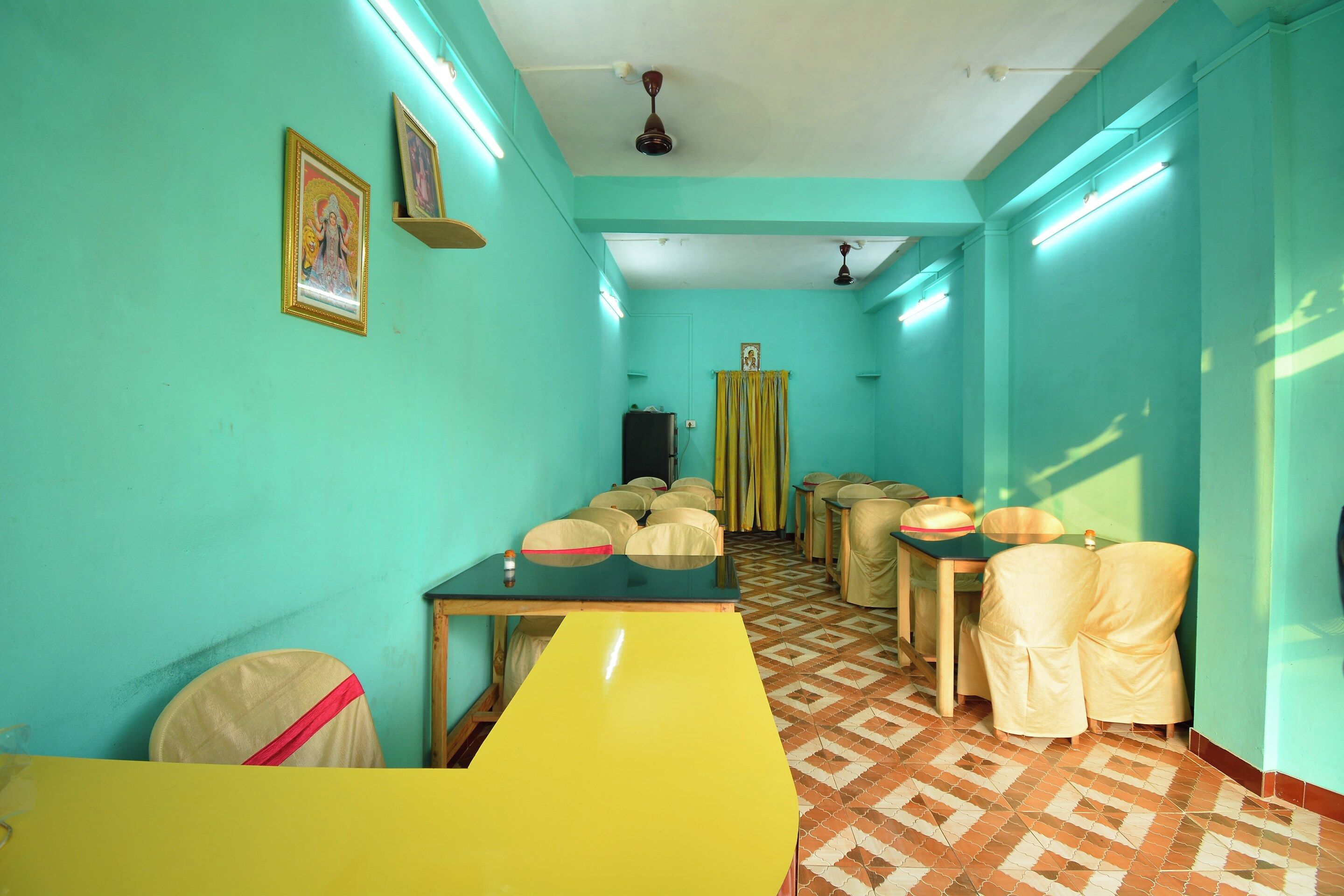 Una habitación con paredes color turquesa, piso estampado y una mesa amarilla. Hay sillas con reposacabezas y un ventilador de techo.