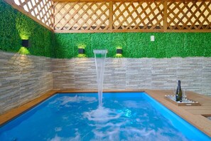 An indoor swimming pool with a waterfall feature, surrounded by stone walls and a wooden lattice ceiling. - Jacuzzi Privado/ Patio Climatizado / TV 75” / A/C (Santo Domingo Este)