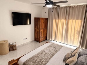 Ein Schlafzimmer mit einem Bett, einem Holzschrank, einem Fernseher, einem Deckenventilator und einem Fenster mit Vorhängen.