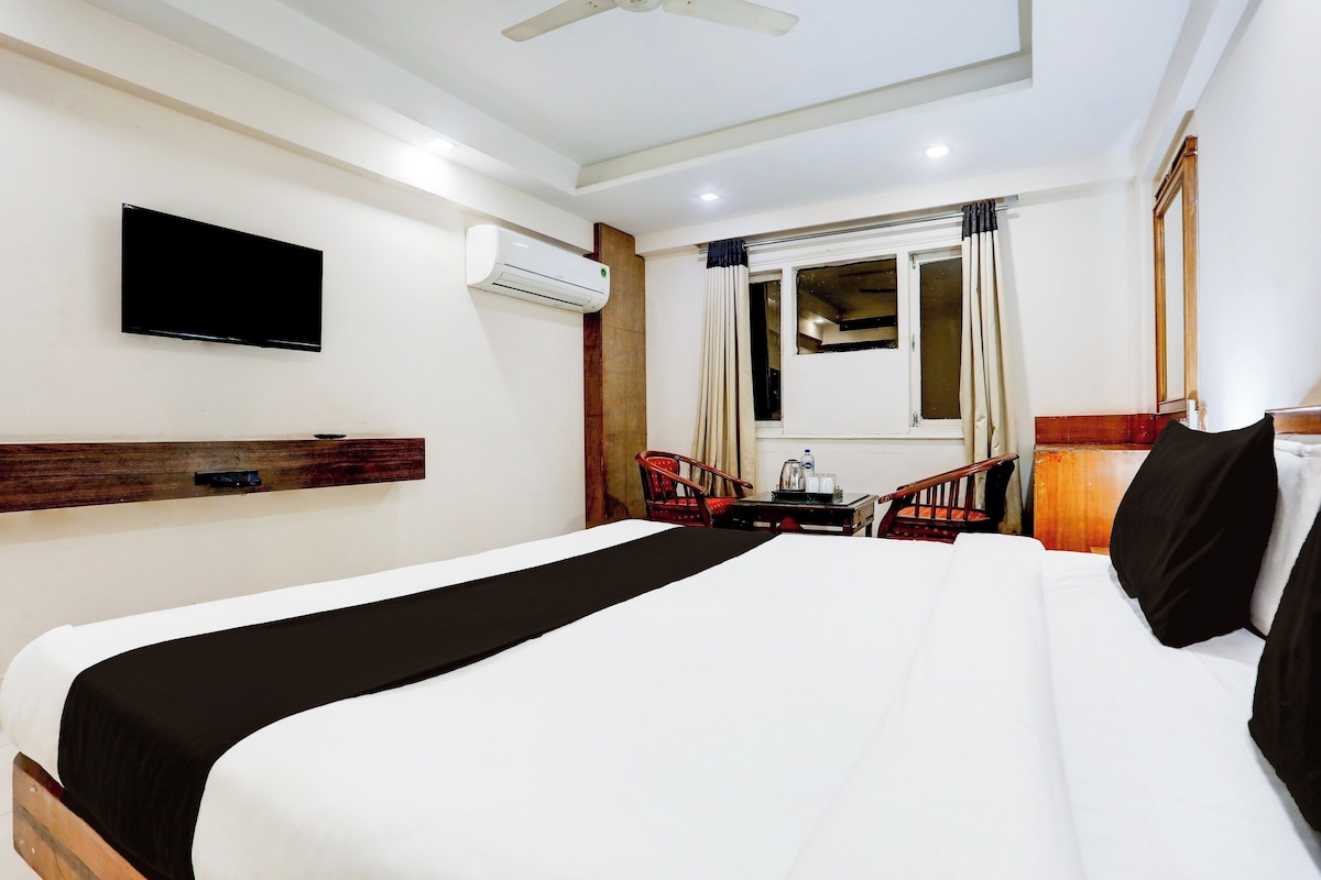 Habitación de hotel con cama, televisor de pantalla plana, equipo de aire acondicionado, ventana con cortinas y una mesita con hervidor.