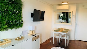 A modern room with a flat-screen TV, a small desk with a lamp, a dining table with chairs, and a green wall decoration.