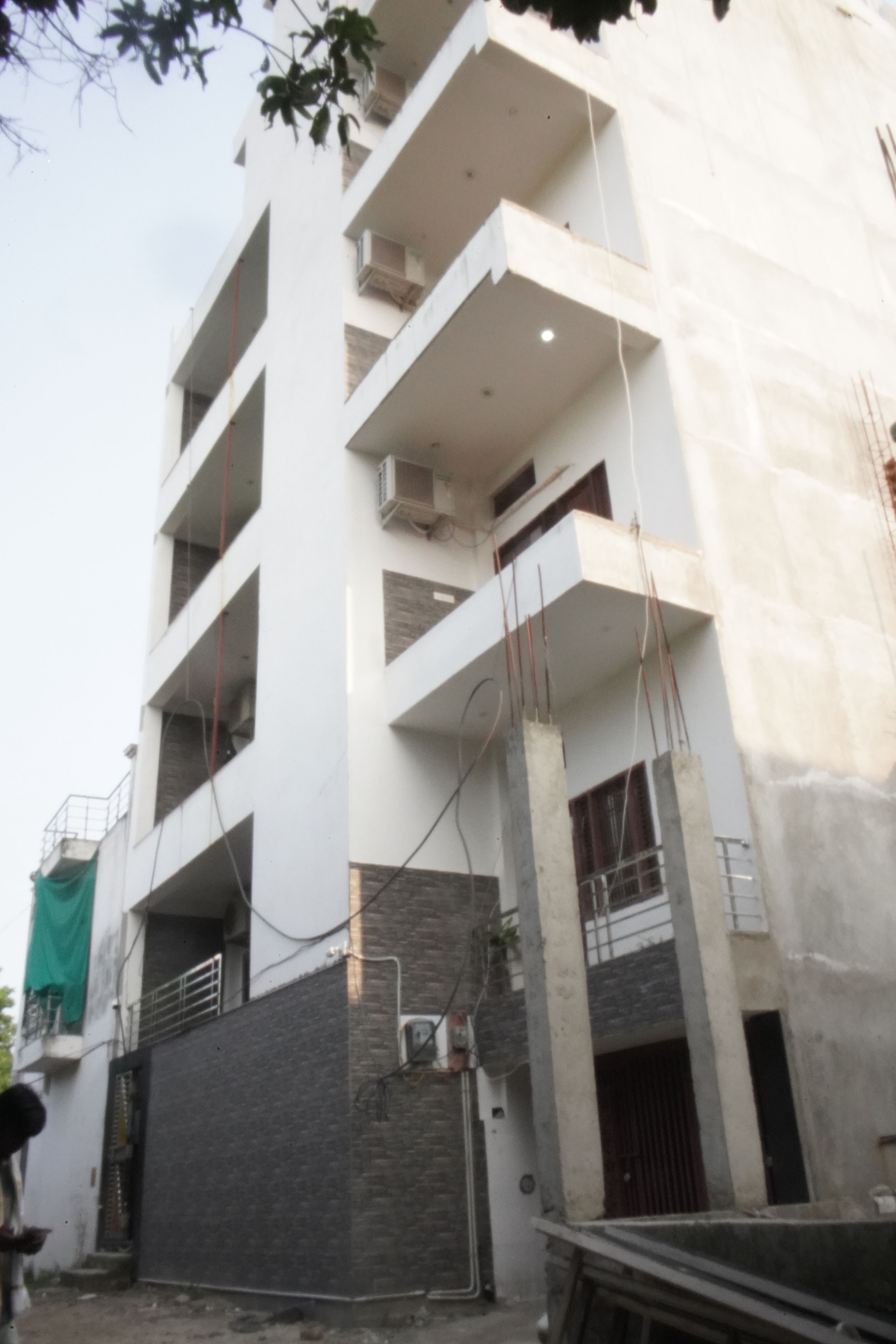 A multi-story building under construction with exposed concrete pillars, a flat roof, and a balcony on the upper floors.