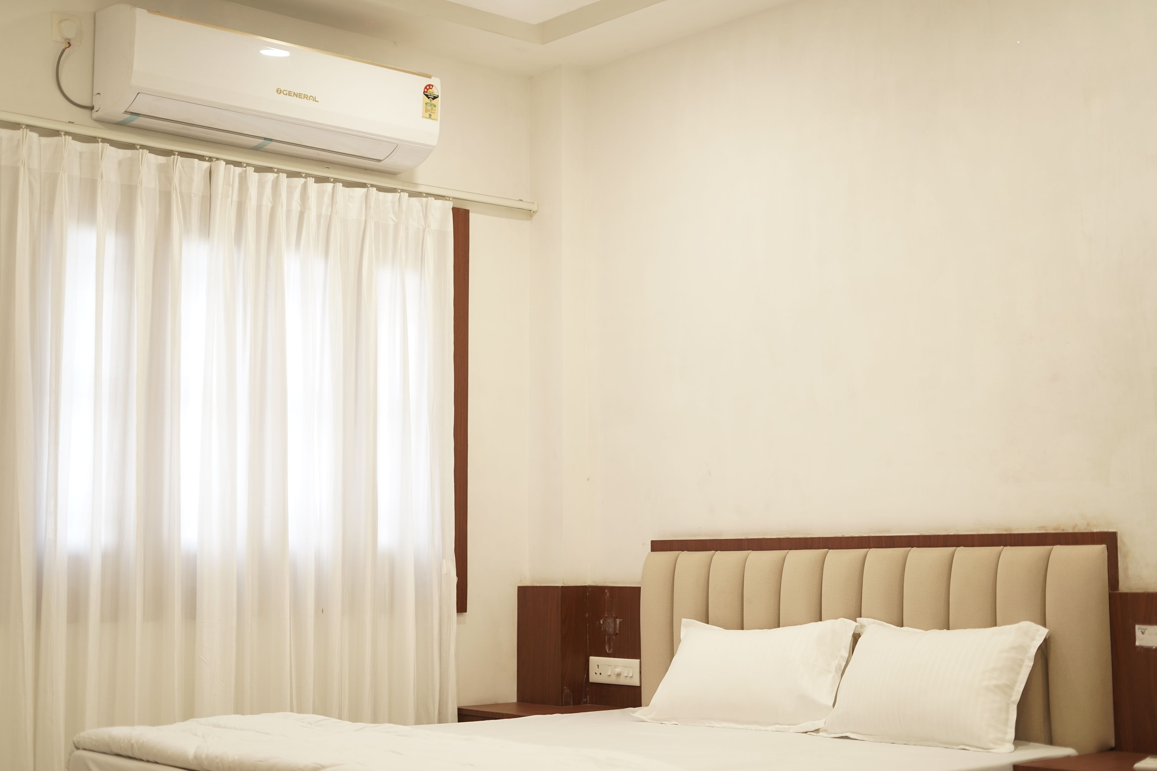 A hotel room with a bed, a nightstand, a wall-mounted air conditioner, and a window with curtains.