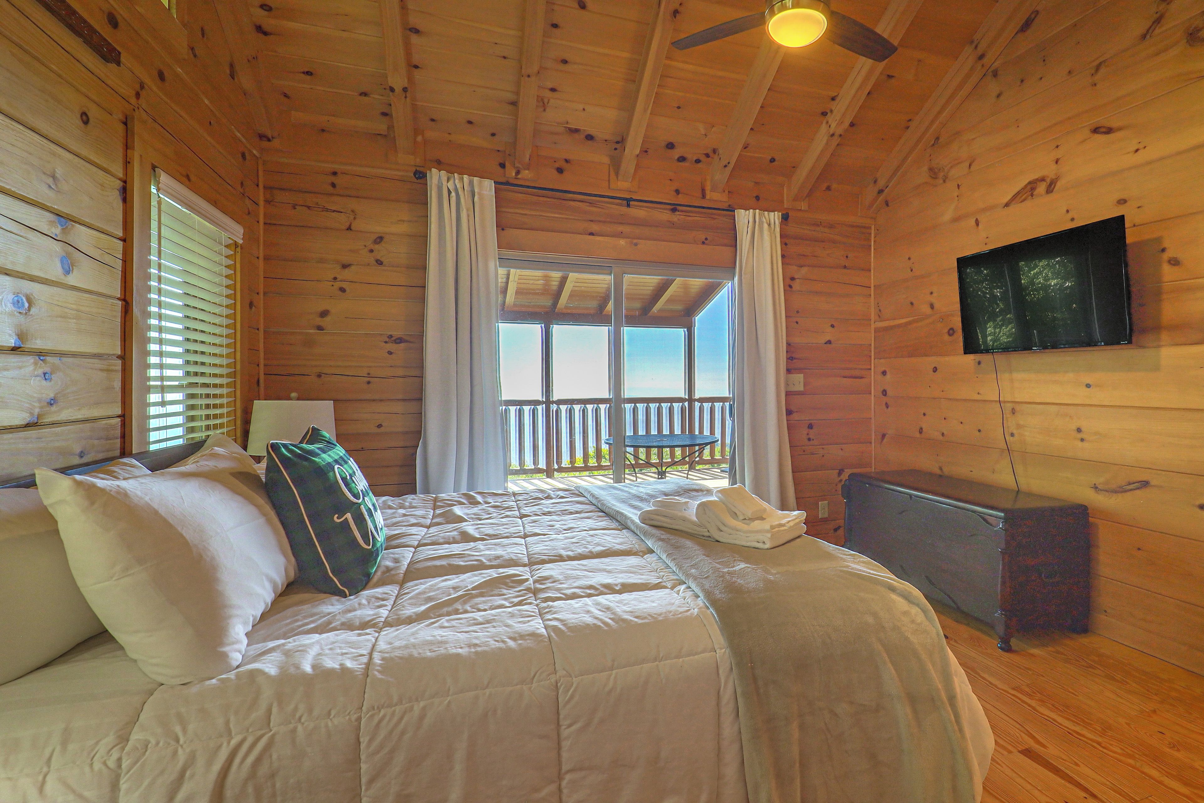 A wooden cabin bedroom with a bed, a television, and a balcony with a view.