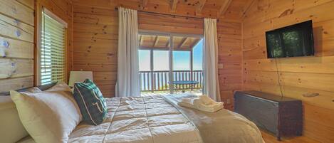 A wooden cabin bedroom with a bed, a television, and a balcony with a view.