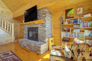 A cozy cabin interior with a stone fireplace, wooden bookshelves, and a staircase. - Hot Tub, Fire Pit, Views & More: Blowing Rock Gem (Blowing Rock)