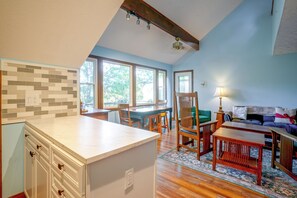 Una cocina con encimera blanca, armarios de madera y salpicadero de azulejos. Hay una zona de comedor con una mesa y sillas de madera, y un sofá al fondo.