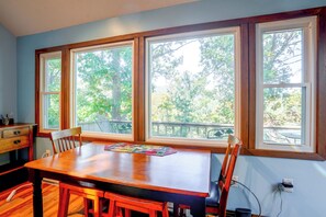 Un área de comedor con mesa y sillas de madera, ventanas amplias con vistas a árboles y un enchufe en la pared.