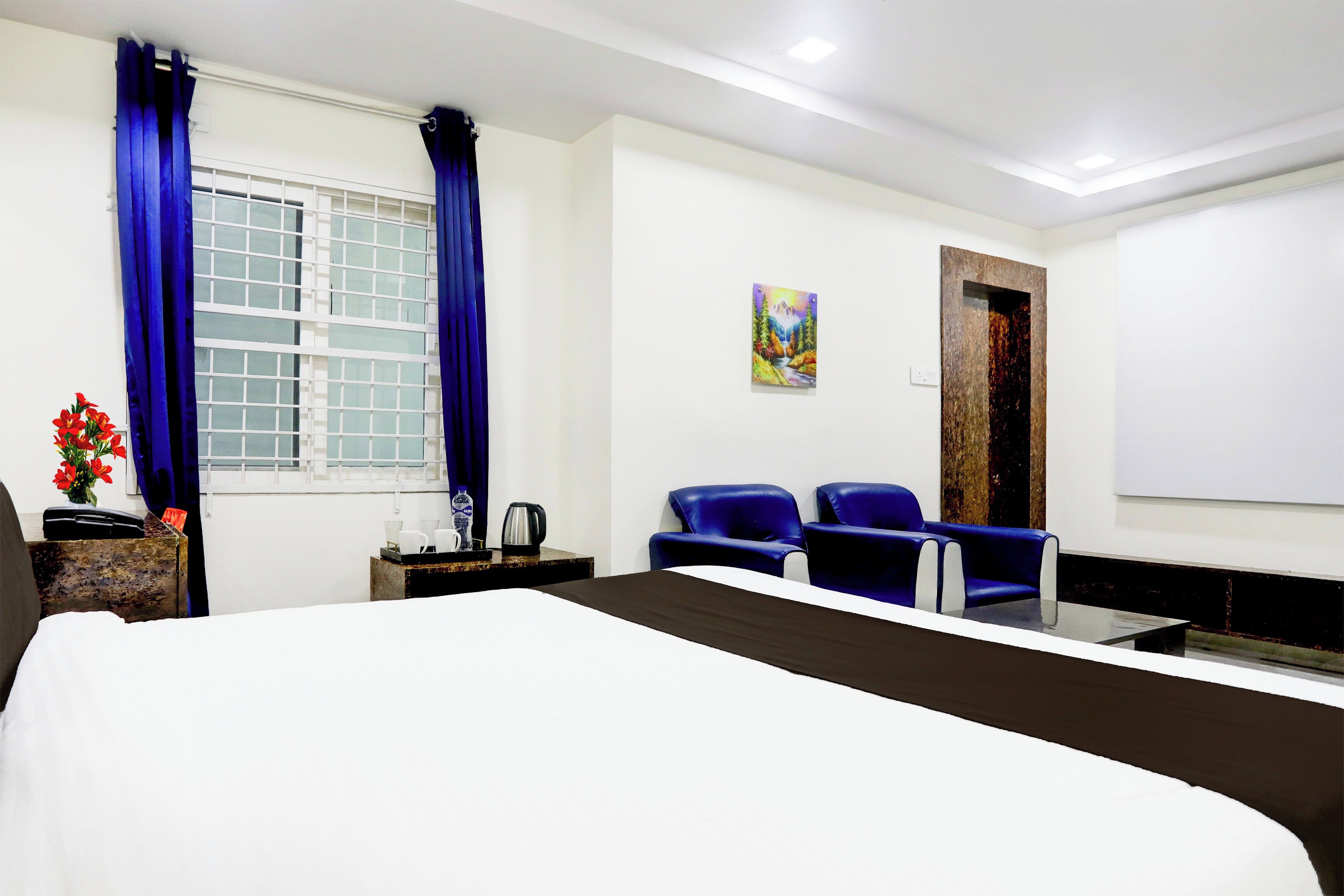 A hotel room with a bed, two blue chairs, a painting on the wall, and a window with curtains.