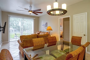 A living room with a glass-top coffee table, wicker chairs, a brown sofa, a ceiling fan, and a door leading to another room. - Resort Perks! Family Condo 4 Mi to Disney World! (Kissimmee)