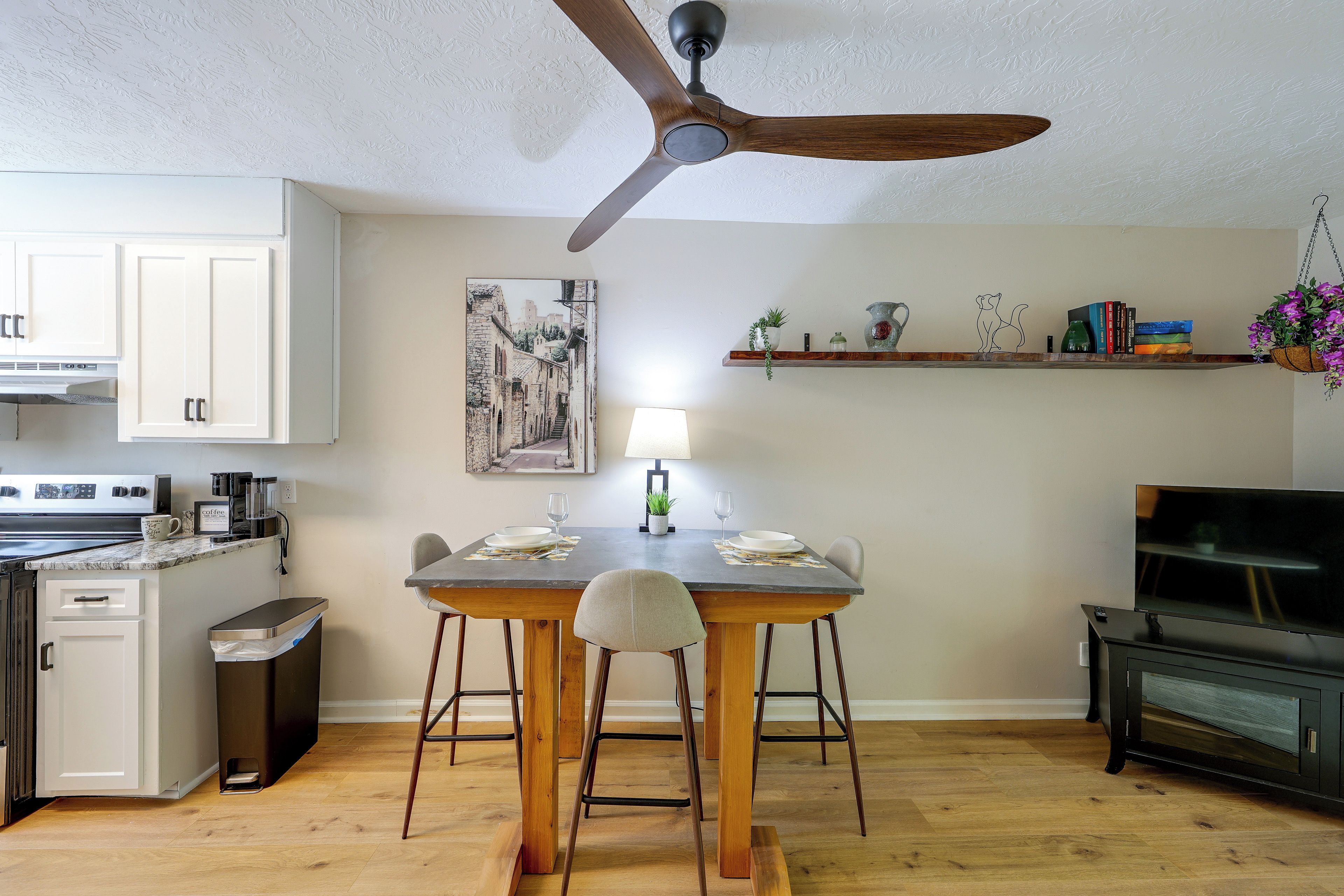 Una cocina moderna con gabinetes blancos, una mesa de comedor con sillas, un ventilador de techo y una pequeña isla con una cafetera.