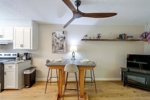 Una cocina moderna con gabinetes blancos, una mesa de comedor con sillas, un ventilador de techo y una pequeña isla con una cafetera.