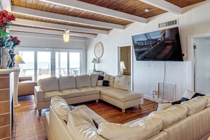 A living room with a sectional sofa, a flat-screen TV mounted on the wall, and a large window overlooking a balcony. - Direct Daytona Beach Access & Views! Spacious Home (Daytona Beach Shores)