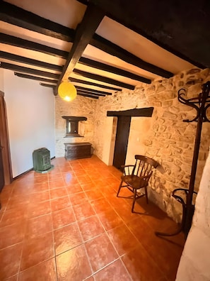 Una habitación con pared de piedra, una silla de madera, un calefactor verde y una lámpara colgante.