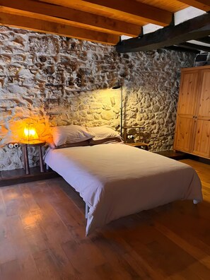Een slaapkamer met een stenen muur, houten vloer, een bed met wit beddengoed, een nachtkastje met een lamp en houten kasten.