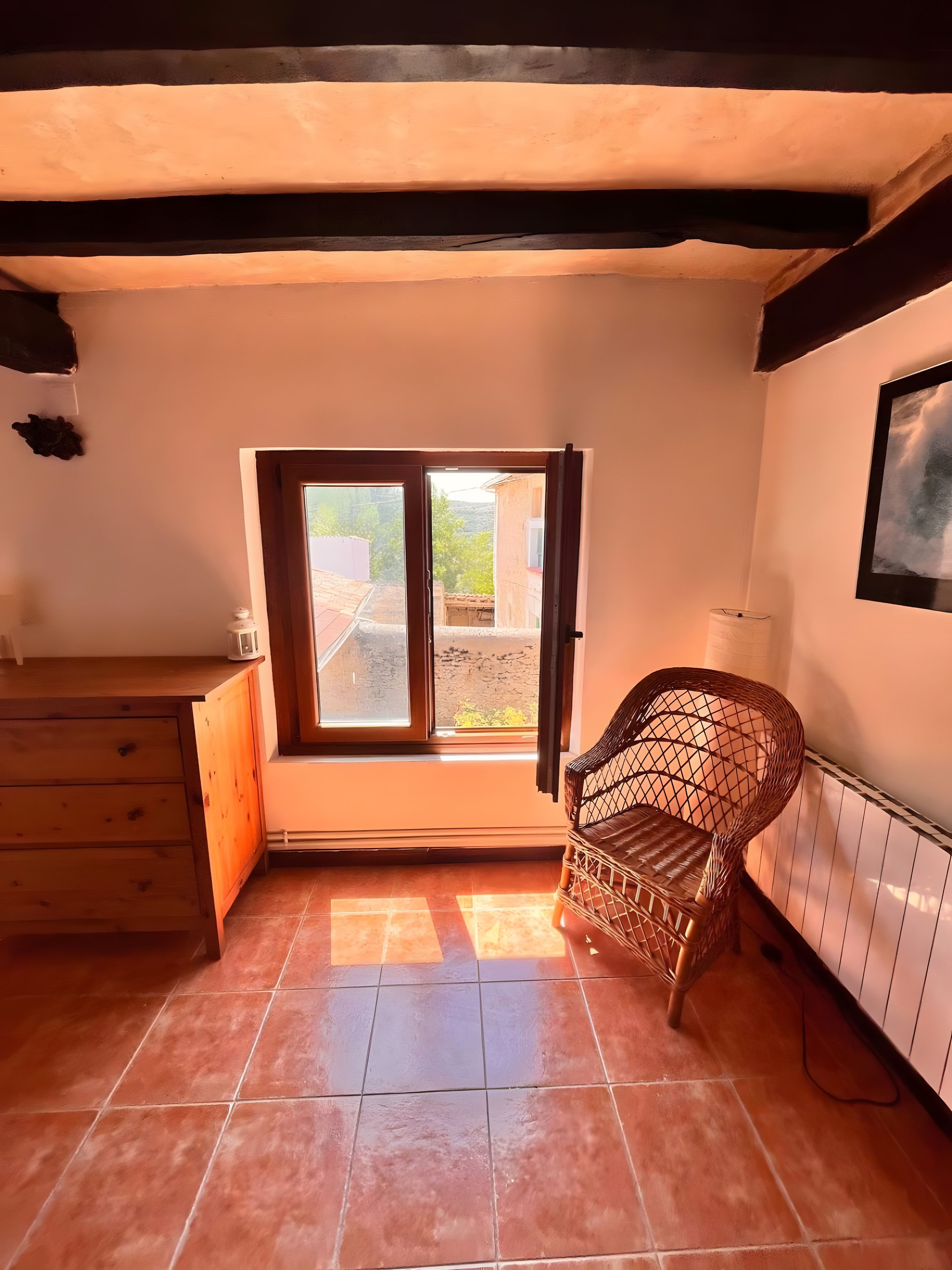Een kamer met een houten kast, een rieten stoel, een raam met uitzicht en een betegelde vloer.