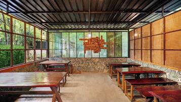 A room with a high ceiling, wooden beams, and large windows. There are multiple tables and benches arranged for seating.