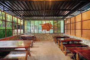 A room with a high ceiling, wooden beams, and large windows. There are multiple tables and benches arranged for seating.