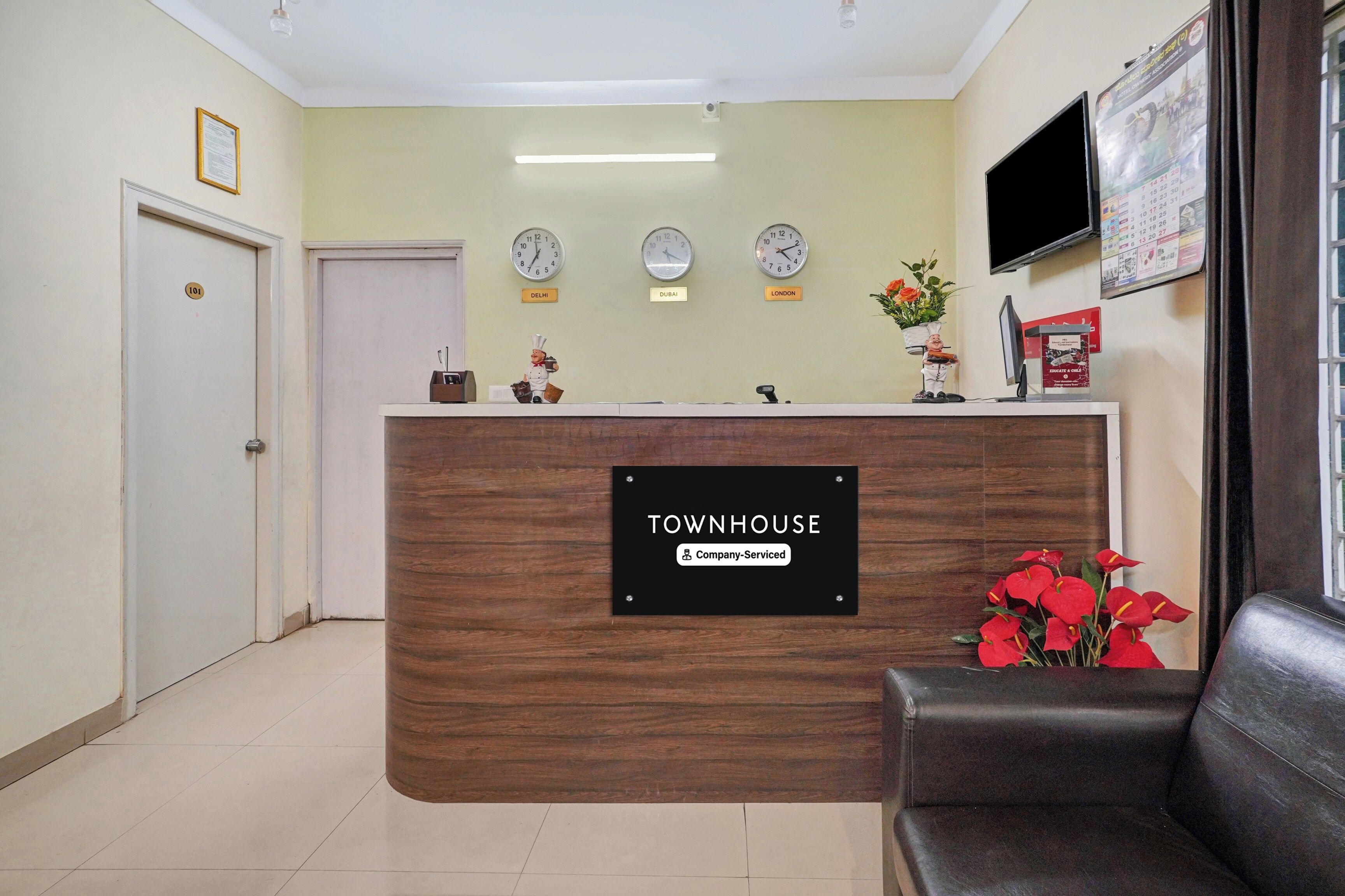 A reception desk with a sign indicating it is company-serviced. There are clocks above the desk showing different time zones, a television mounted on the wall, and a door labeled '101'.