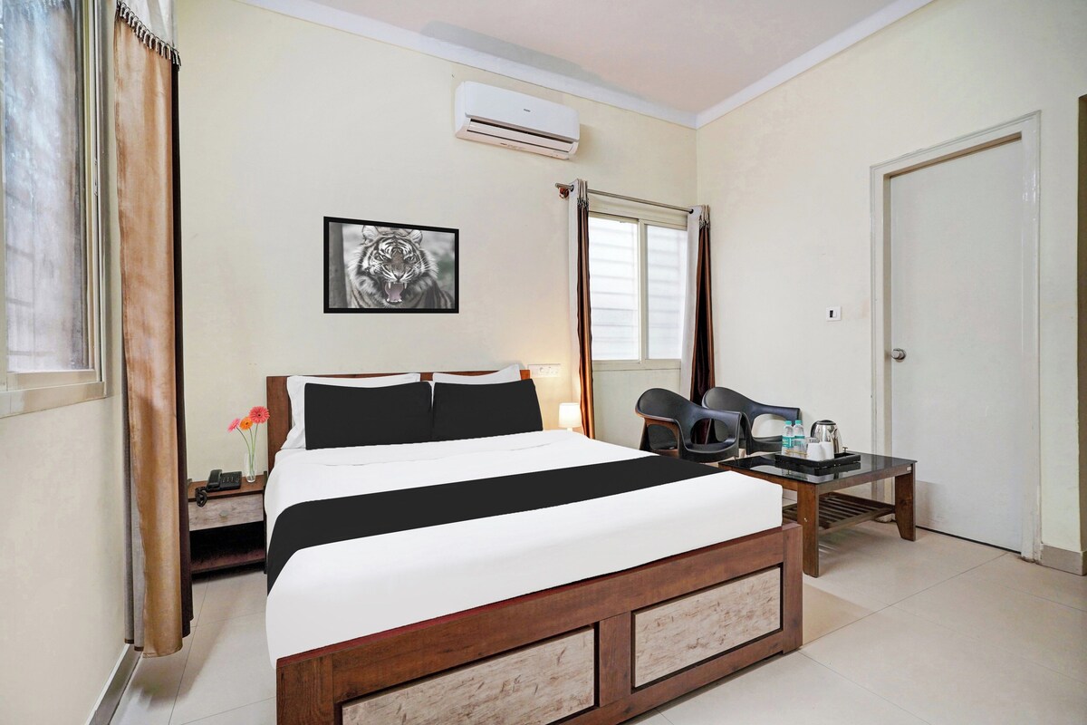 A hotel room with a bed, a bedside table with a phone and a vase of flowers, a chair, a table with a kettle and water bottles, an air conditioning unit, and a door.