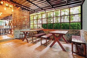 An indoor dining area with wooden tables and benches, a patterned wall, and large windows with a view of greenery. - Townhouse Hebbal Mysore (Shrirangapattana)