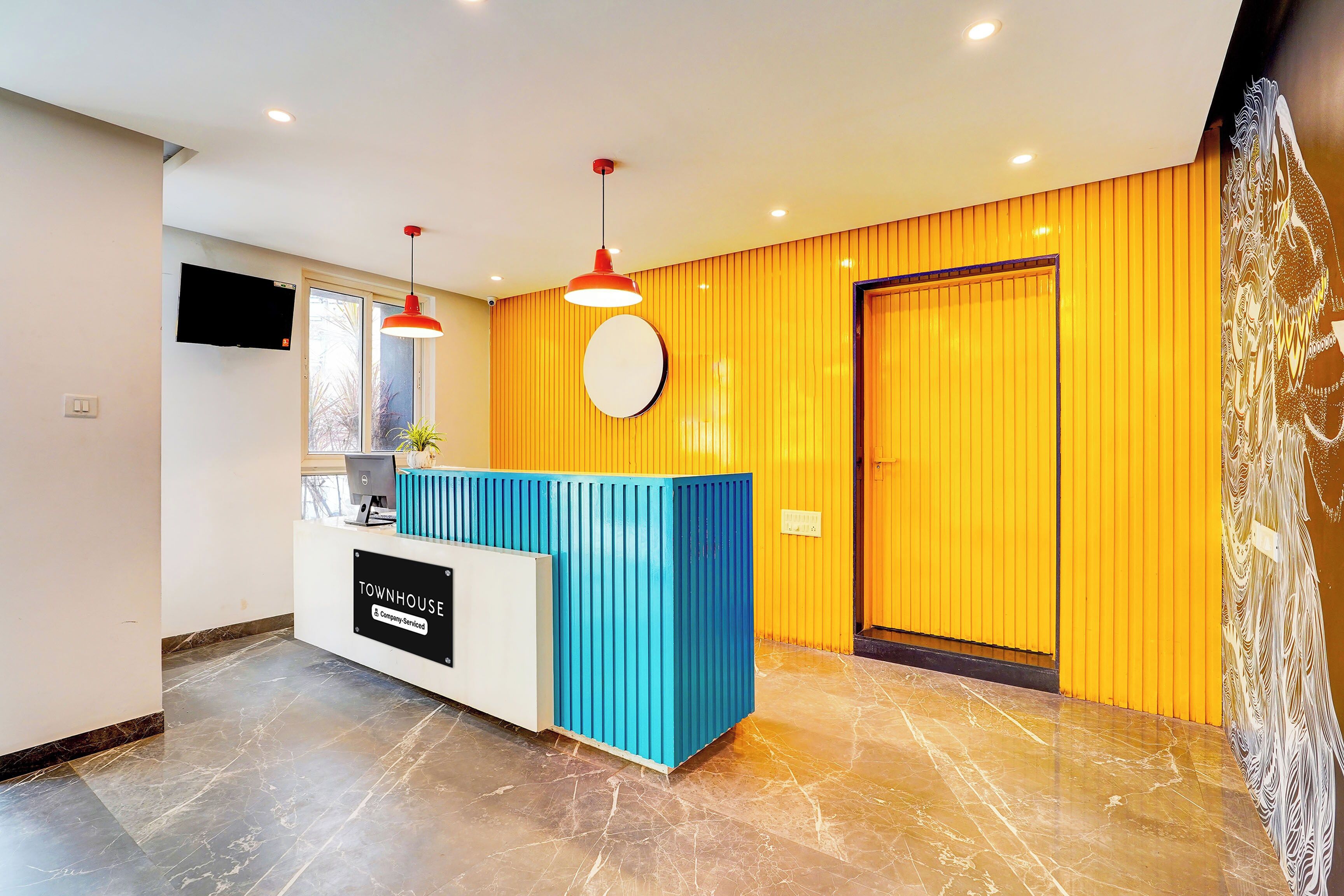 A reception desk with a sign indicating it's a company-serviced location, a flat-screen TV mounted on the wall, and a door with a decorative pattern.