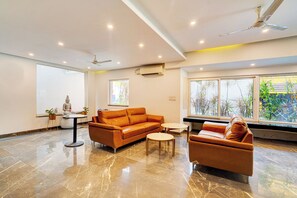 En moderne stue med sofa, lænestol og et lille bord. Loftventilator, indbygget belysning og et stort vindue med udsigt til grønne omgivelser.