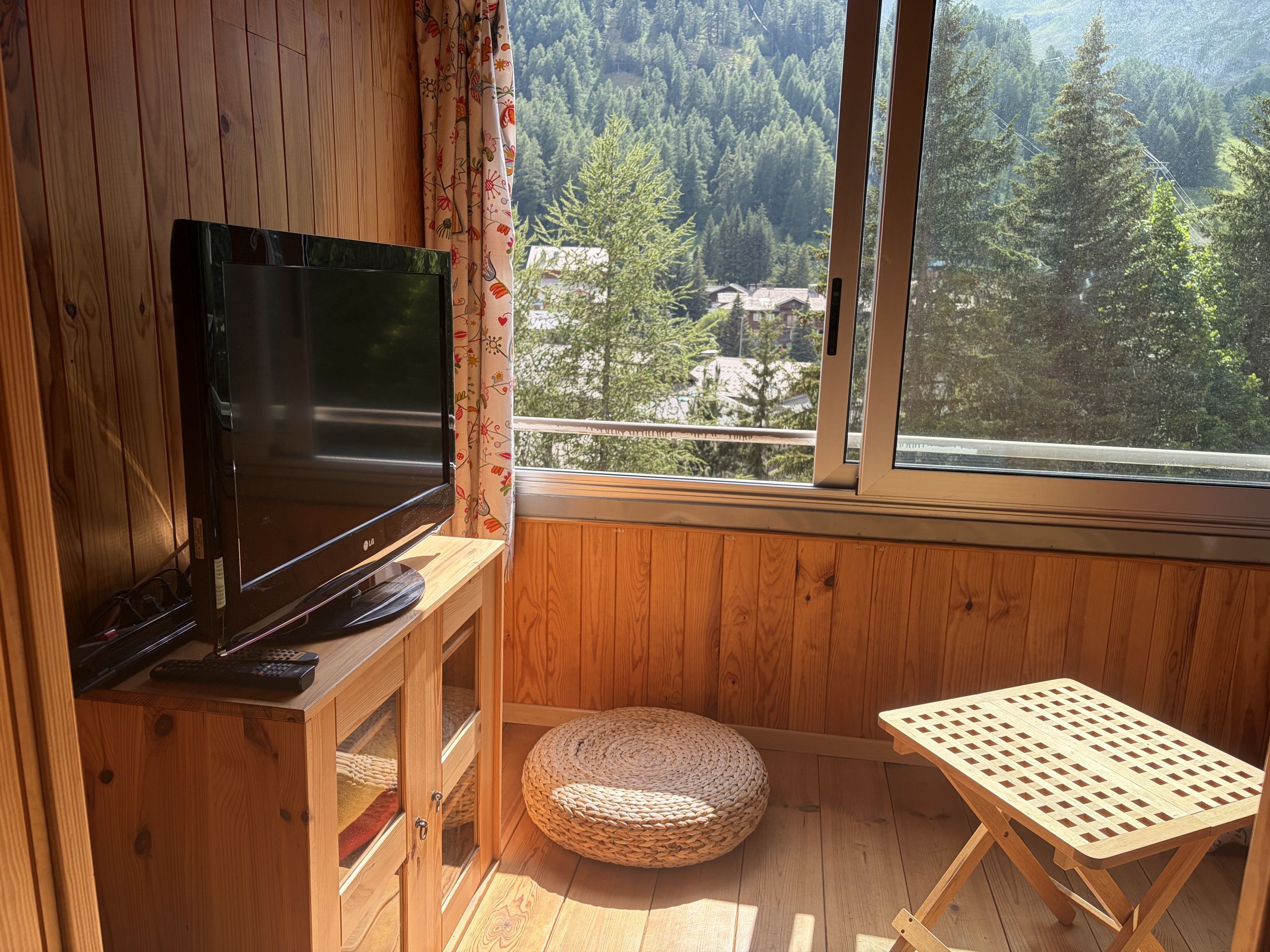 Une pièce en bois avec un téléviseur posé sur un meuble, un petit pouf rond en osier, une table en bois et une fenêtre donnant sur une zone boisée.