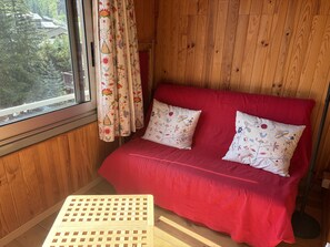 Ein Zimmer mit Holzwänden, einem roten Sofa, gemusterten Vorhängen und einem kleinen Holzschemel.