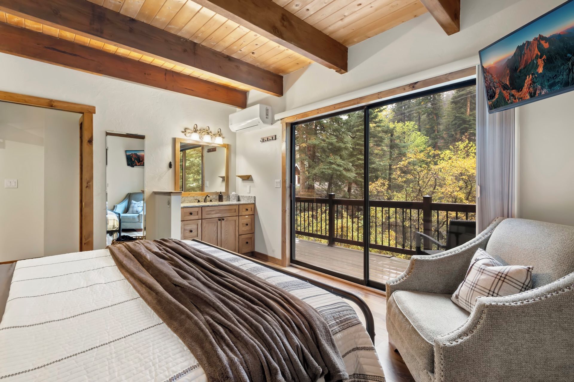 A bedroom with a large bed, a chair, a bathroom with a mirror, and a balcony with a view of trees.