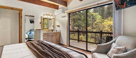 A bedroom with a large bed, a chair, a bathroom with a mirror, and a balcony with a view of trees.