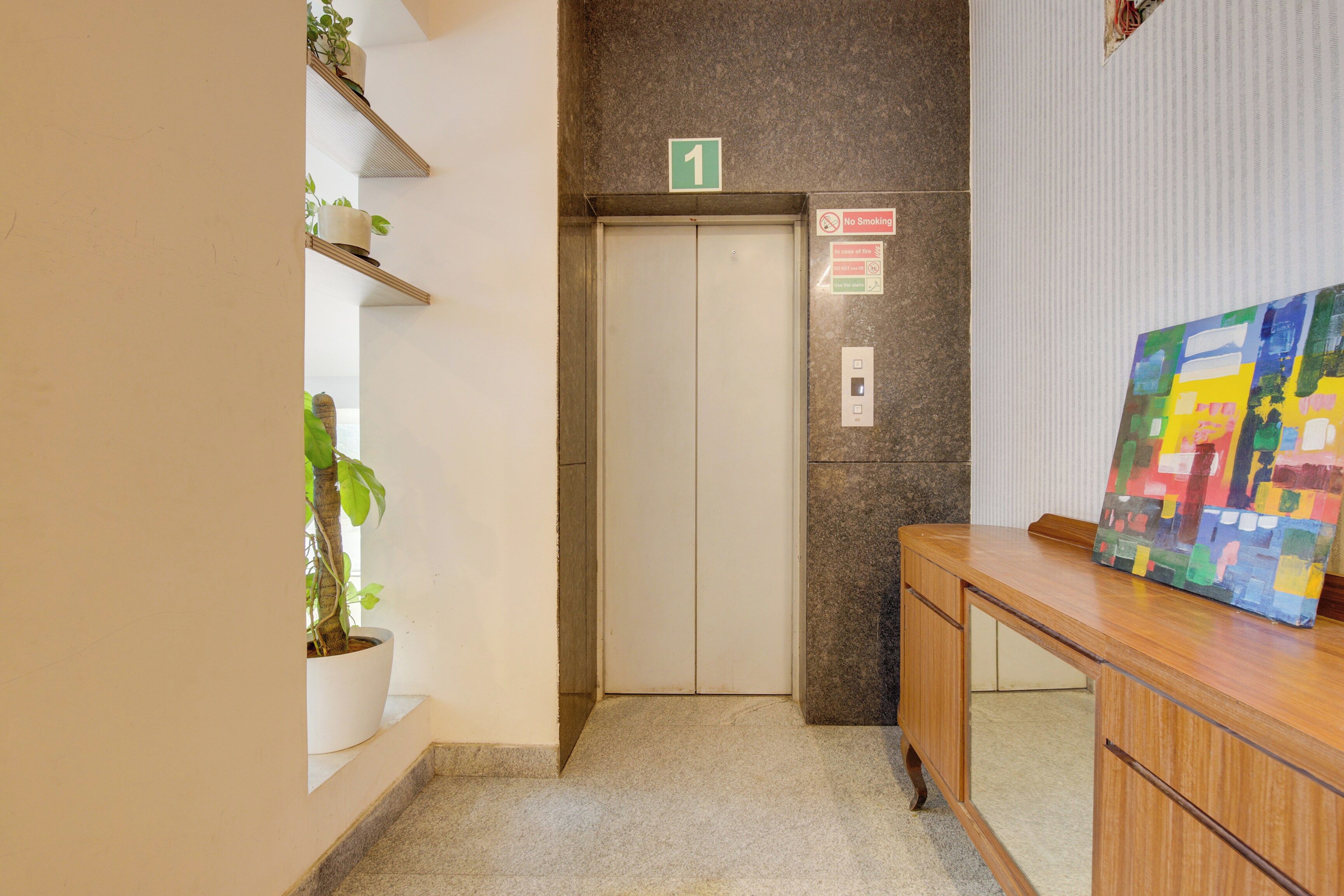 Un ascensor con un cartel de 'No Fumar', un indicador verde con el número '1' y un cuadro sobre un escritorio de madera.