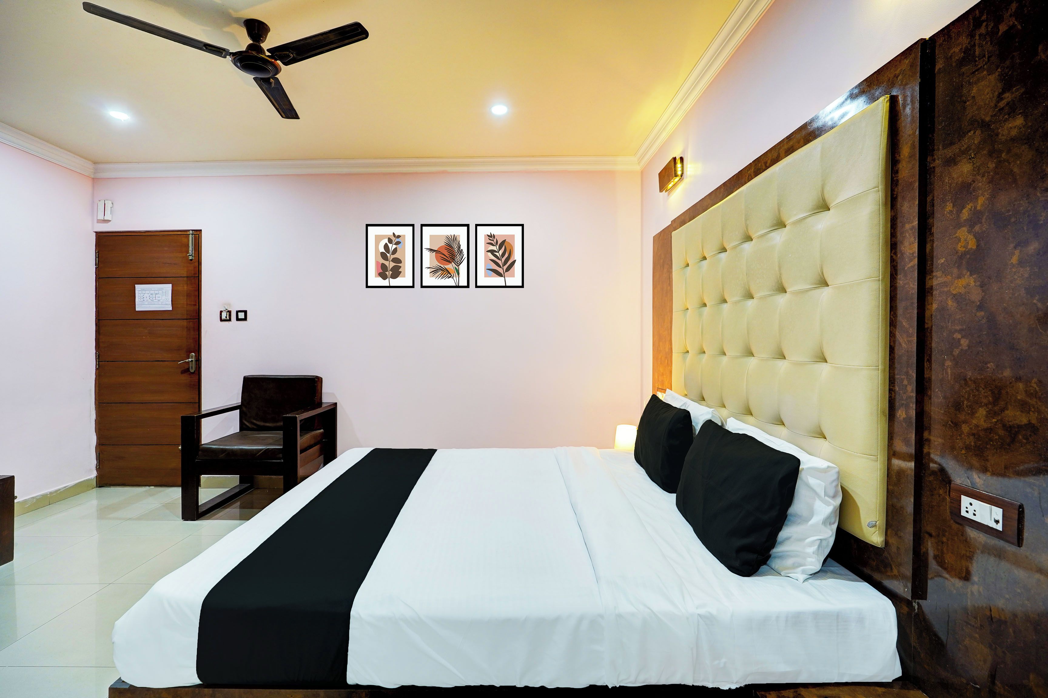 A hotel room with a bed, a chair, a ceiling fan, and three framed artworks on the wall.