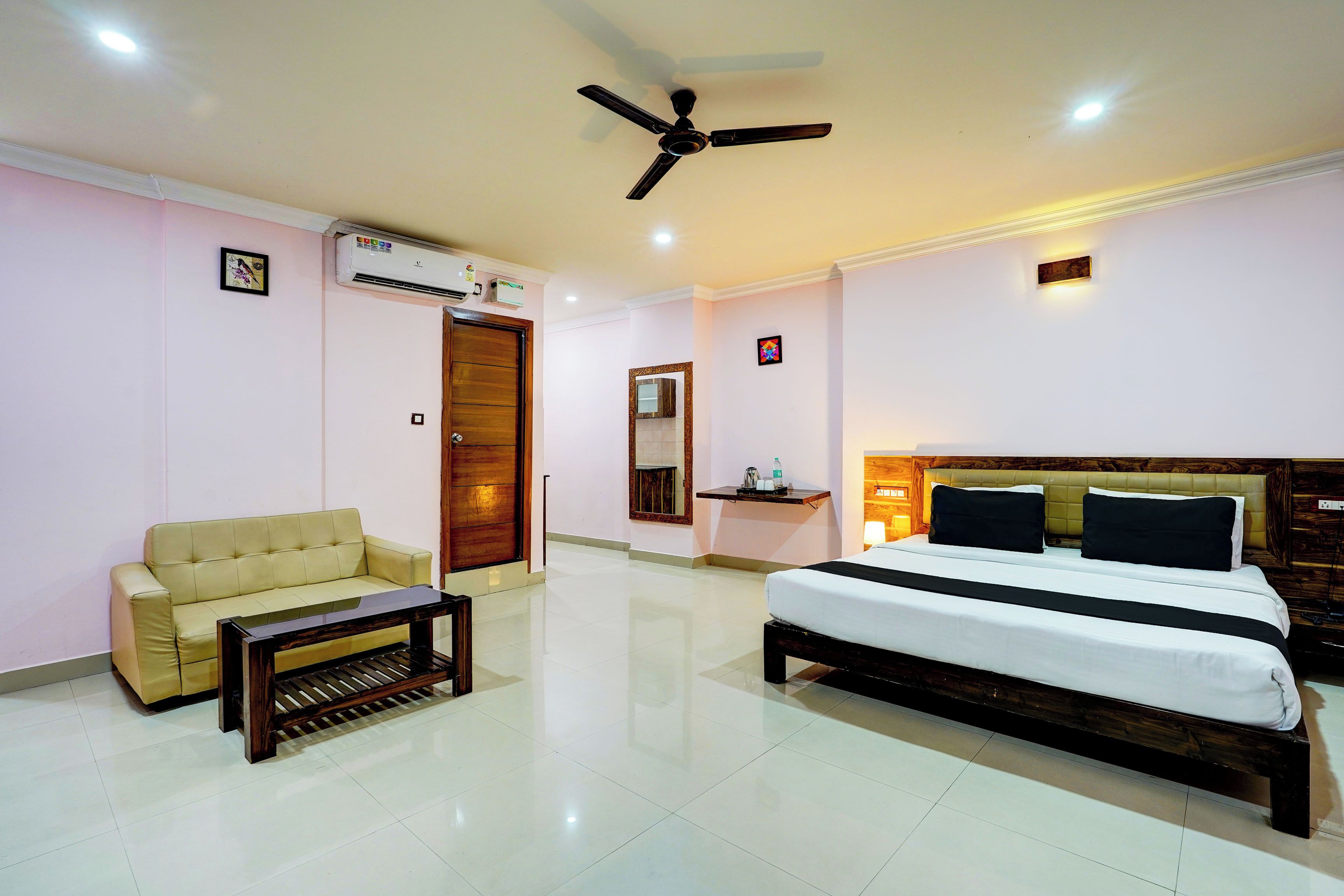 A hotel room with a bed, a sofa, a small table, an air conditioning unit, a ceiling fan, and a mirror.