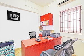 A hotel room with a red desk, a computer, a telephone, a chair, a bed with patterned bedding, a window with bars, and a sign indicating it's a company-serviced hotel. - Hotel O Sholinganallur Near Anjaneya (Chennai)