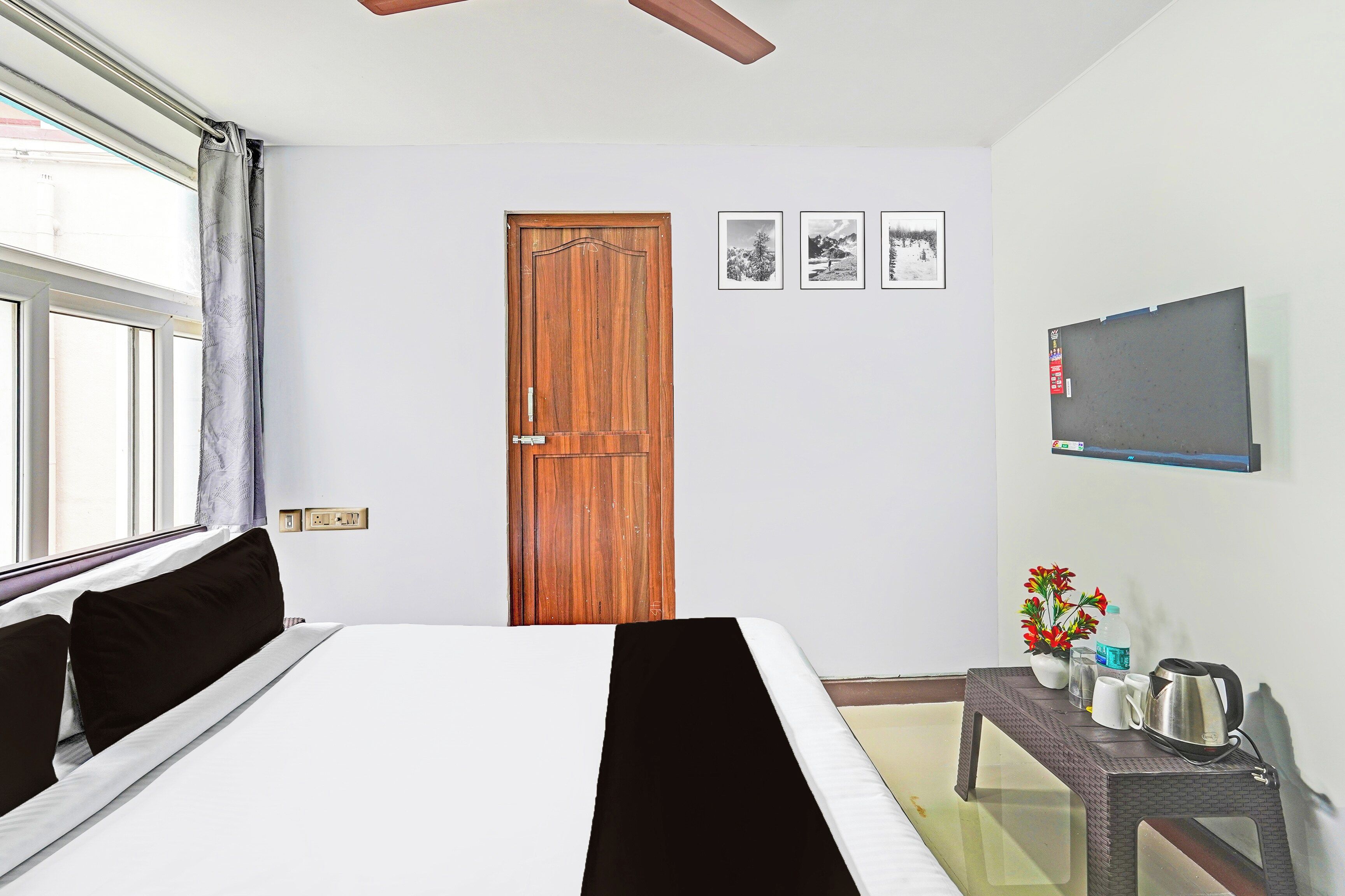 A hotel room with a bed, a wooden door, a wall-mounted TV, and a small table with a kettle and cups.