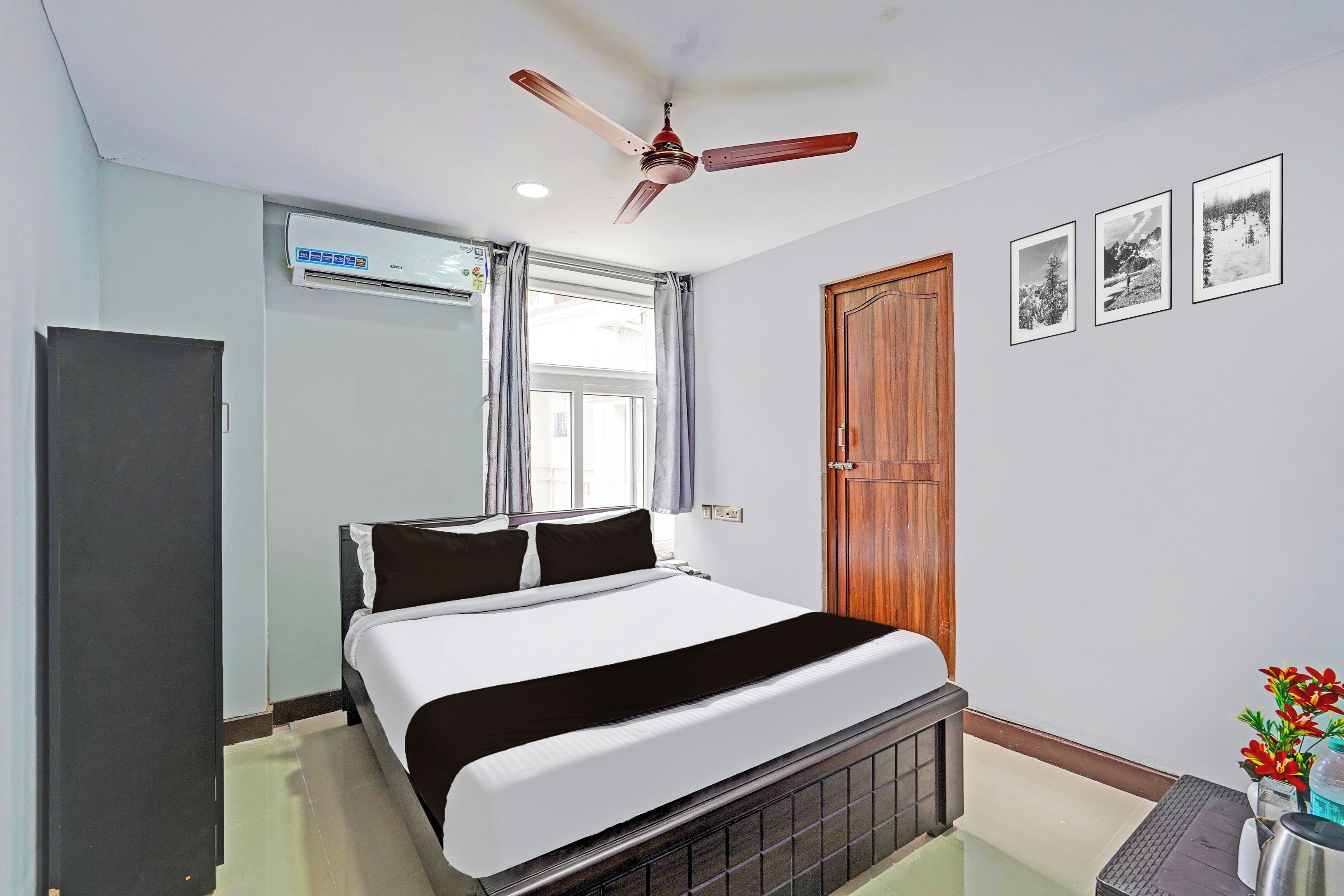 Une chambre d’hôtel avec un lit, un ventilateur de plafond, une fenêtre avec des rideaux, une porte en bois et des tableaux encadrés accrochés au mur.
