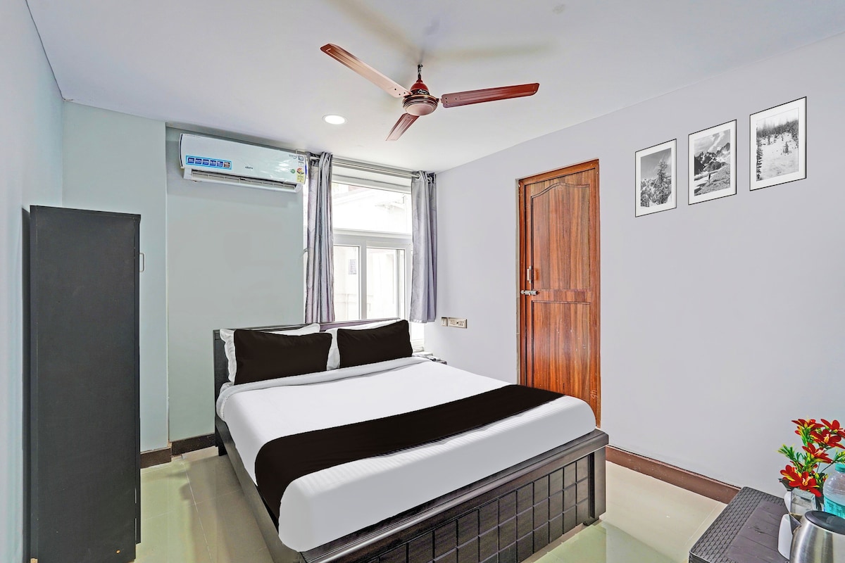 A hotel room with a bed, a ceiling fan, a window with curtains, a wooden door, and framed pictures on the wall.