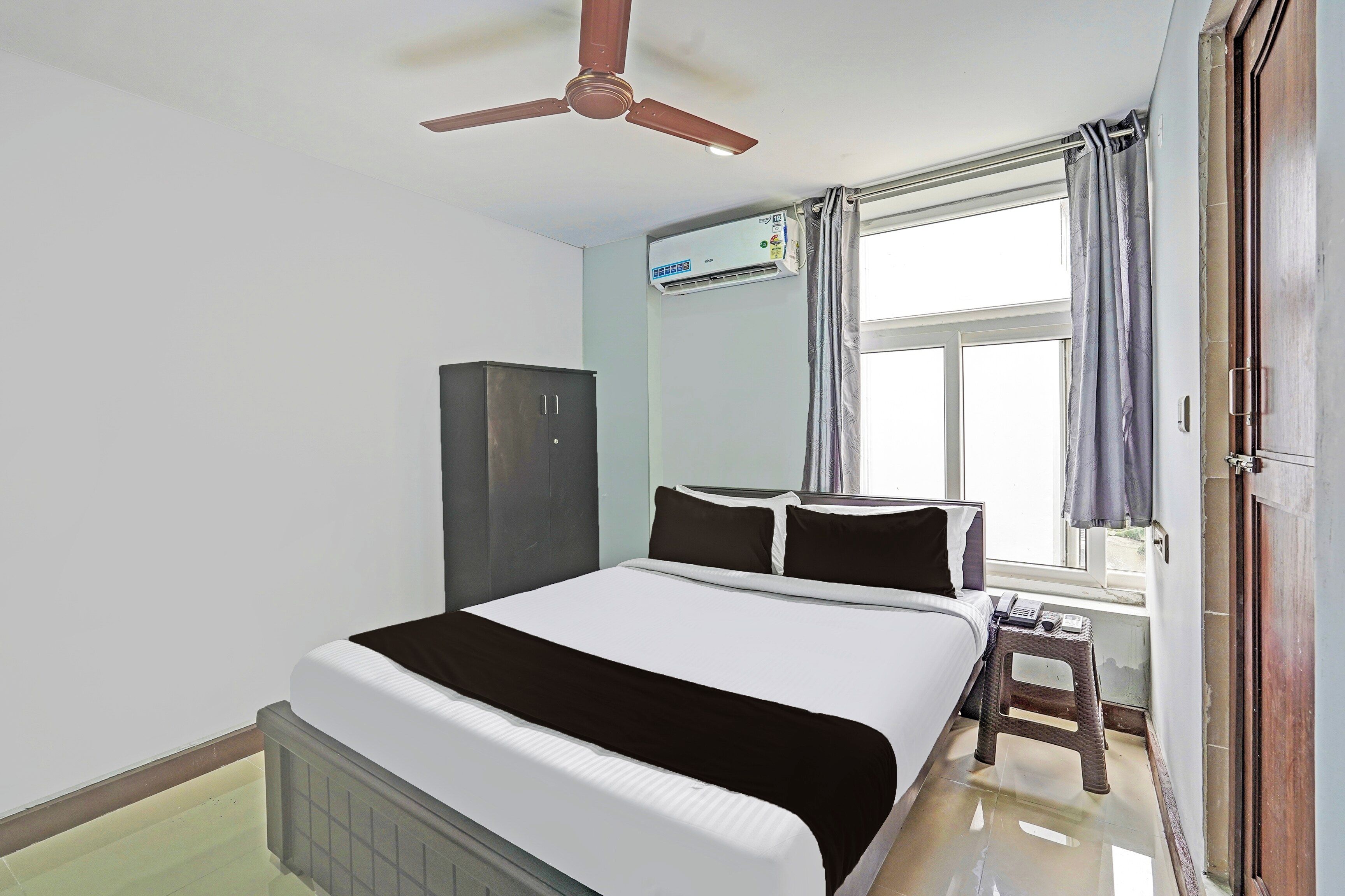 Une chambre d’hôtel comprenant un lit, une armoire, un ventilateur de plafond, un appareil de climatisation, une fenêtre avec des rideaux et un petit tabouret.