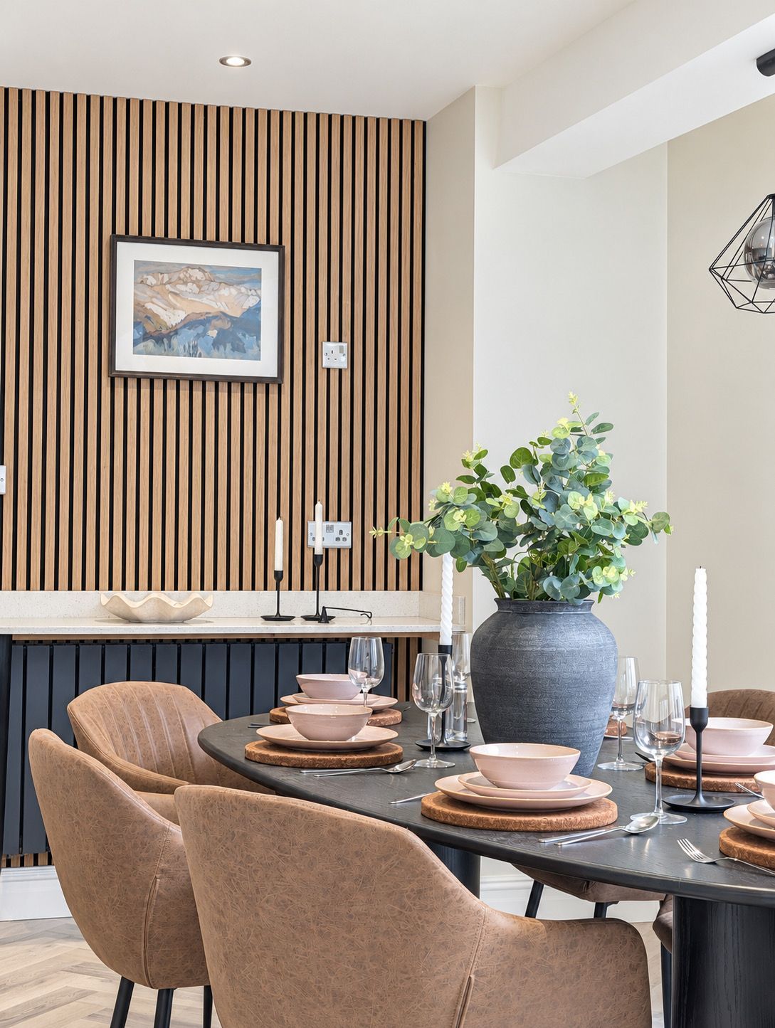 Ein moderner Essbereich mit einer Wandpaneele aus Holz, einem gerahmten Landschaftsbild, einer großen Topfpflanze und einem Tisch, gedeckt für vier Personen, mit rosa Tellern und Gläsern.