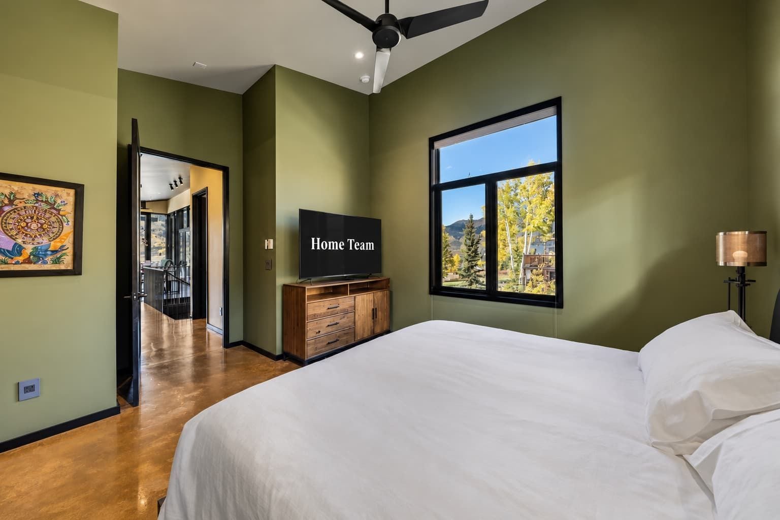 A bedroom with a bed, a television on a wooden cabinet, a window with a view of trees, and a ceiling fan.