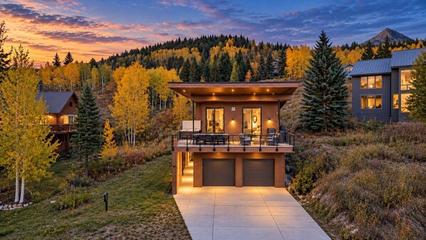 Una casa moderna de dos pisos con balcón, garaje y vista a una montaña boscosa.