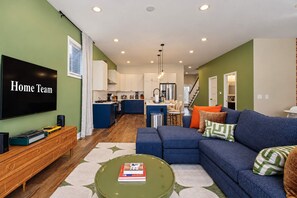 A modern living room with a large flat-screen TV, a blue sofa, and a coffee table with books. - Retro Retreat Hot Tub Firepit Near Lucas Oil (Indianapolis)
