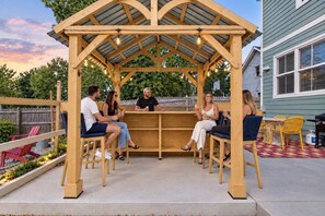 A wooden pergola with string lights, seating, and a basketball hoop in a backyard setting. - Retro Retreat Hot Tub Firepit Near Lucas Oil (Indianapolis)
